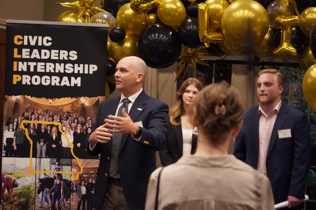 Chadis Follis addresses an audience while standing in front of a CLIP banner and a black and gold balloon arch.