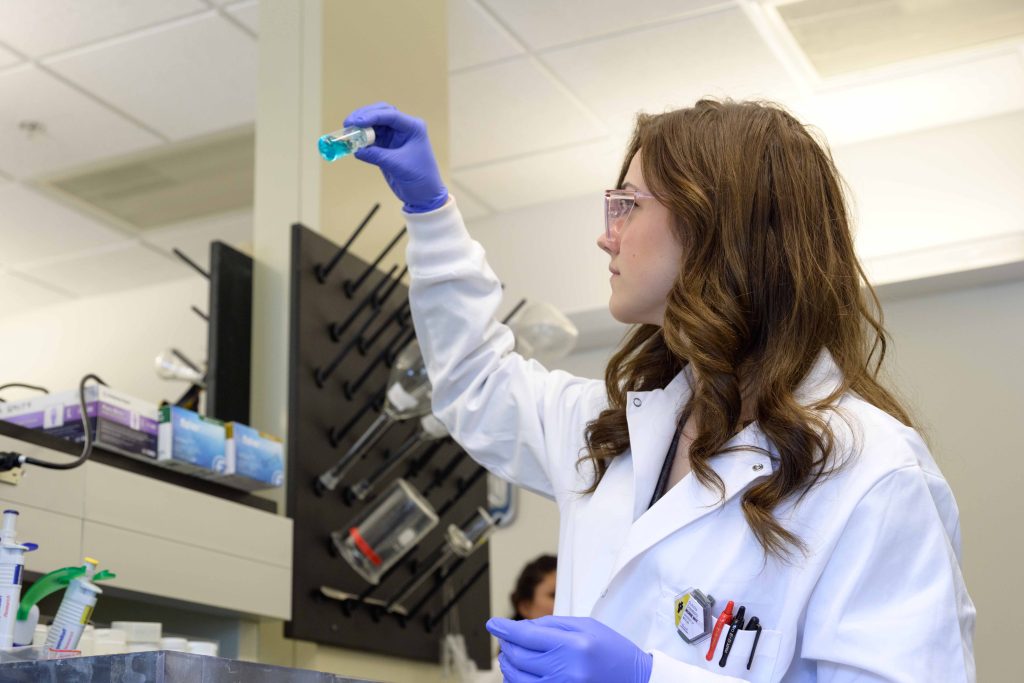 Claudia Chambers, Ph.D standing in lab with protective eyewear, white lab coat holding up a container of blue liquid  analyzing it.