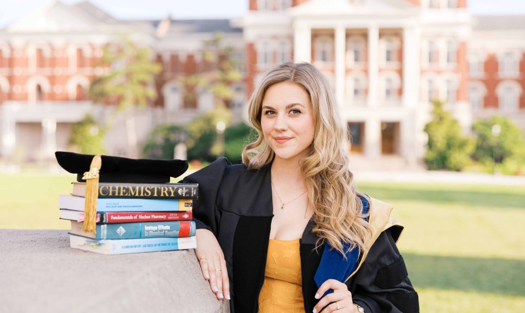 Claudia Chamber, PhD wearing doctoral gown, poses for portrait with Jesse Hall in the background. Her doctoral tam is placed on a stack of textbooks with gold 2025 and tassel.