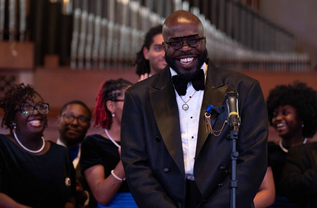 G. Preston Wilson, smiling on stage in front of a microphone in a black tuxedo with the Fisk Jubilee Singers behind him.