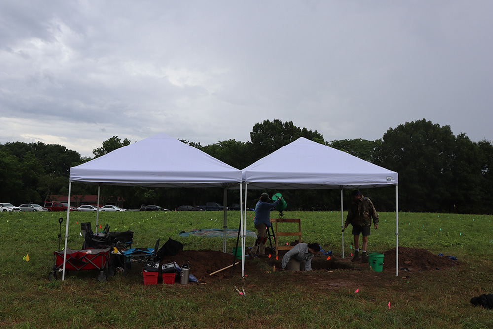 Students working under the tents in the middle of the field digging excavation units at the Bothwell Lodge State Historic Site. 